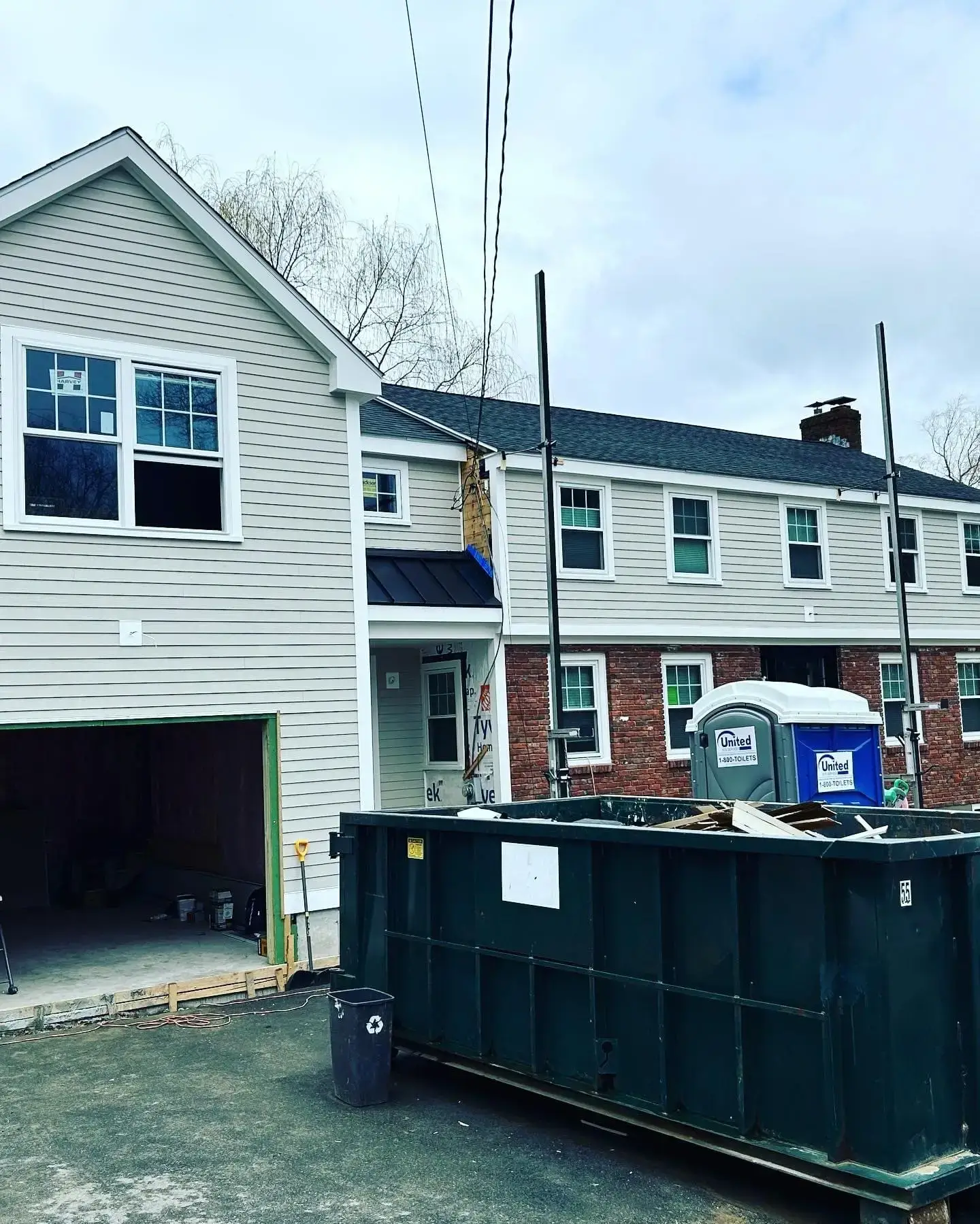 Construction site of a home renovation in South Hamilton, MA, featuring a partially completed exterior with new siding, open garage, construction dumpster, and portable toilets, illustrating Bradford Construction Management's whole-home renovation services.