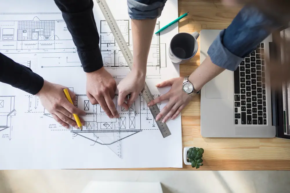 Hands collaborating on architectural blueprints with a ruler and laptop, emphasizing design-build processes in construction.