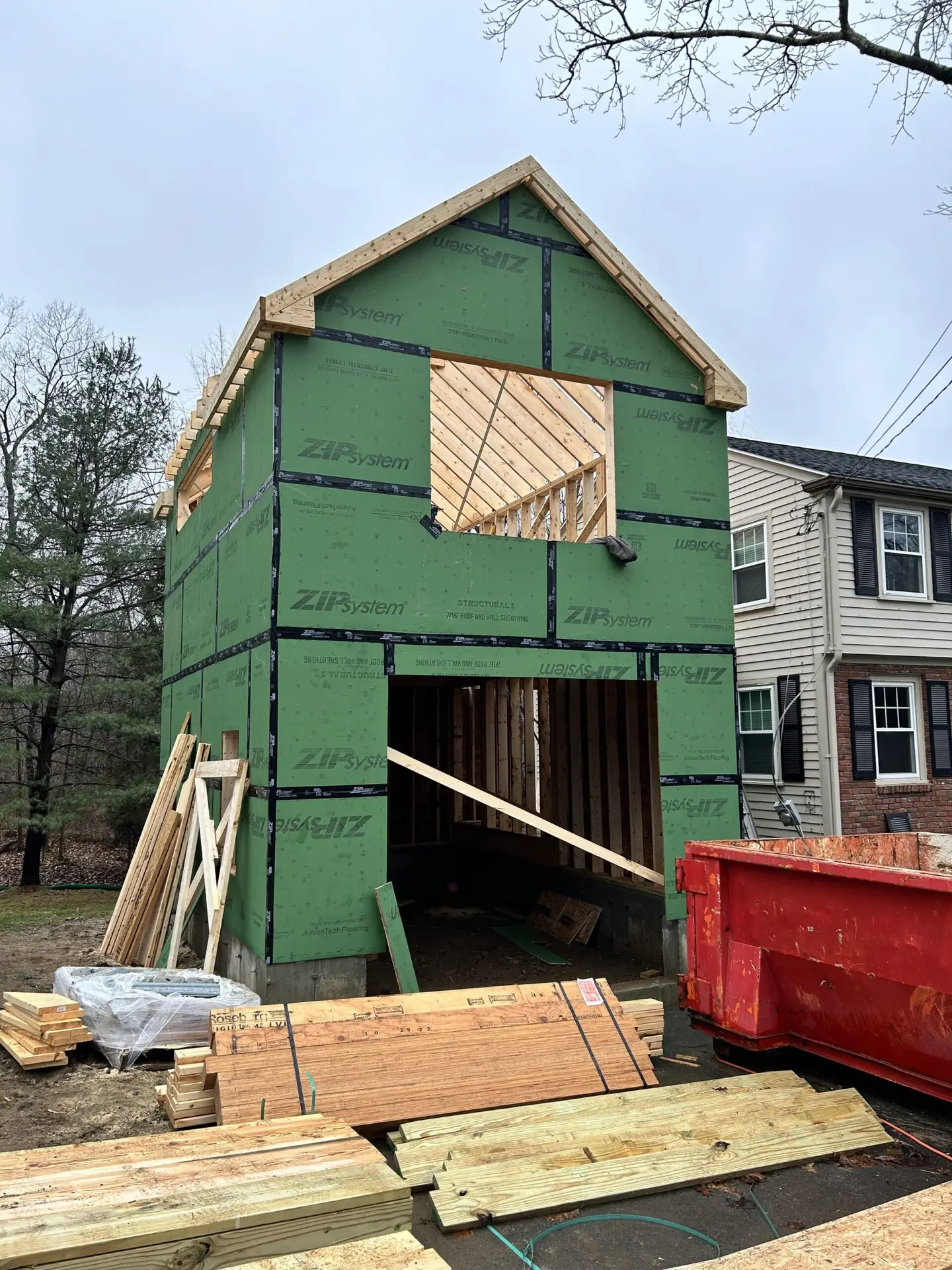 Custom home under construction in Wakefield, MA, featuring ZIP system sheathing, framed roof, and surrounding materials including lumber and a red dumpster.