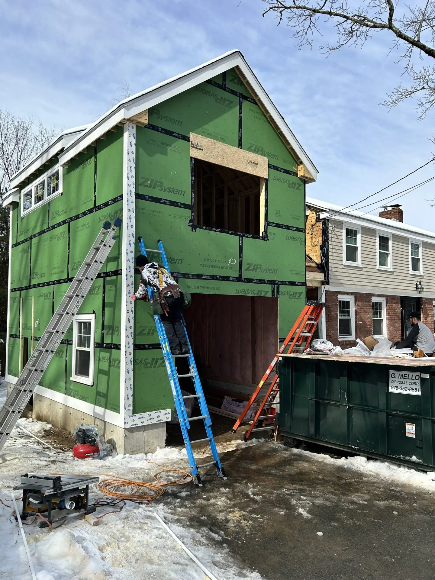 Construction site featuring a green-framed house under renovation, workers on ladders installing exterior sheathing, with tools and a dumpster nearby, showcasing Bradford Construction Management's whole-home renovation services in South Hamilton, MA.