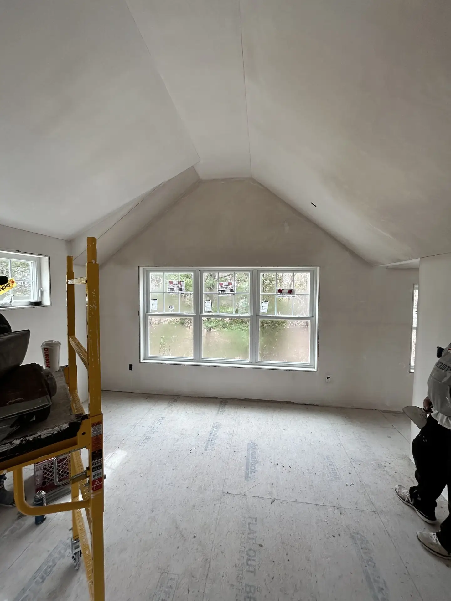 Interior view of a partially finished room with vaulted ceilings, large windows, and a construction worker applying finishing touches, showcasing Bradford Construction Management's renovation work in South Hamilton, MA.
