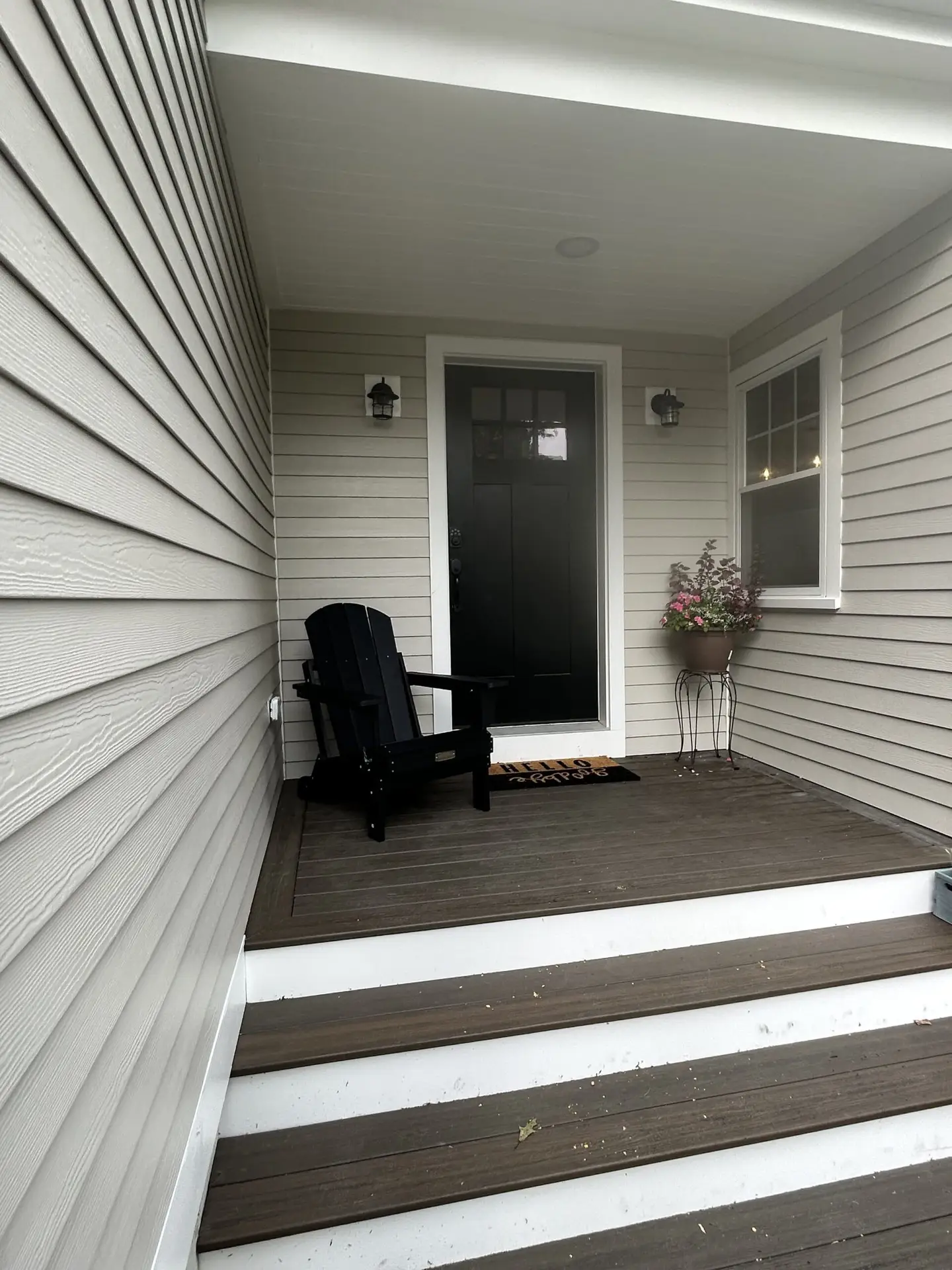 Front porch of a renovated home in South Hamilton, MA, featuring a black Adirondack chair, a black front door with glass panels, decorative wall lights, and a potted plant, highlighting quality craftsmanship and inviting outdoor space.