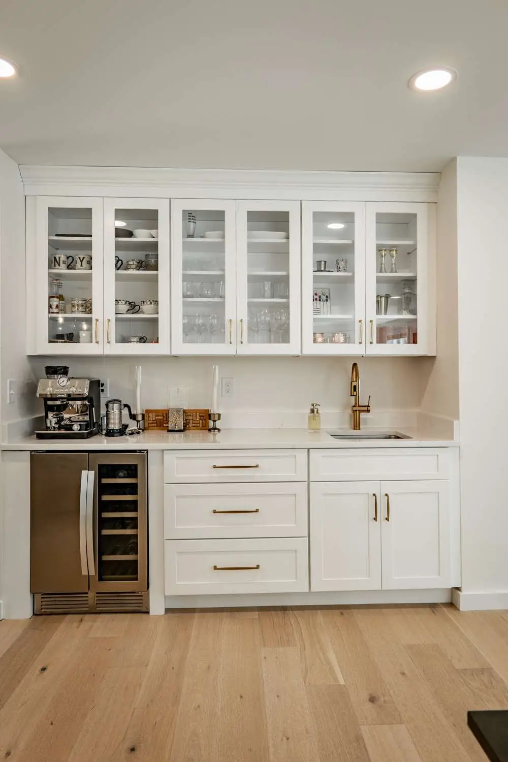 Custom home kitchen design featuring white cabinetry, glass-front upper cabinets displaying glassware, stainless steel appliances, and a gold faucet, highlighting modern aesthetics and functionality in Exeter, NH.