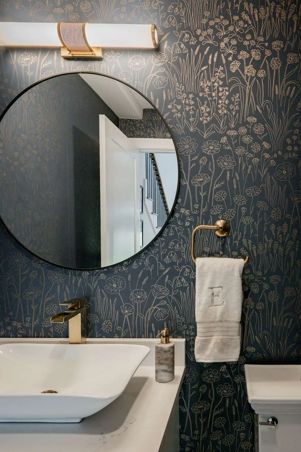 Modern bathroom design featuring a round mirror, stylish light fixture, floral-patterned wallpaper, and elegant sink with gold accents, showcasing custom home aesthetics in Exeter, NH.