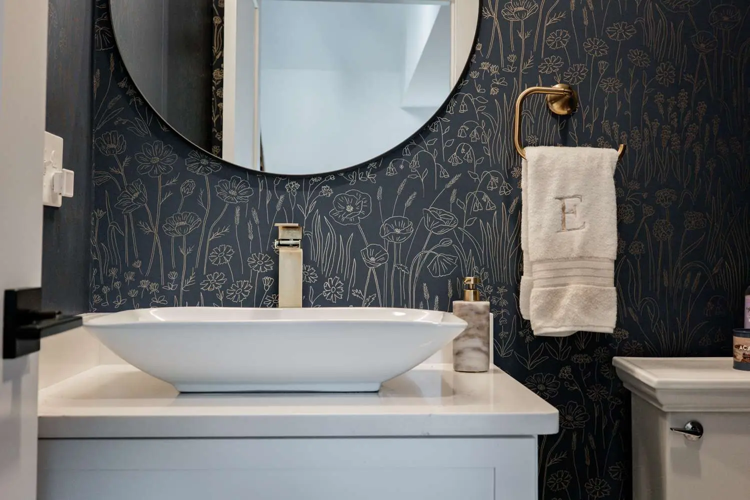 Modern bathroom design featuring a white vessel sink, gold faucet, decorative floral wallpaper, and a towel with an embroidered letter E.