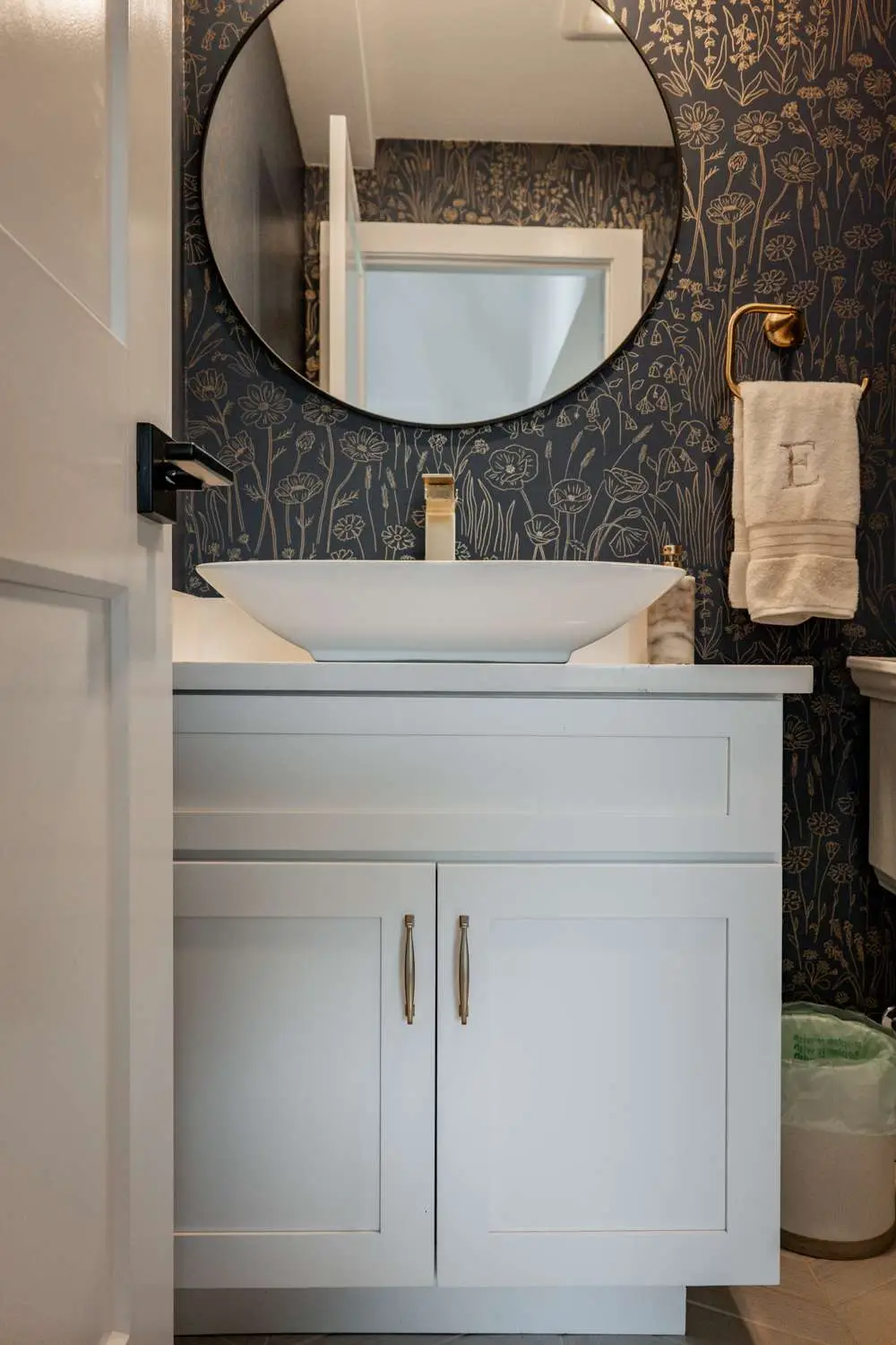 Modern bathroom vanity with white cabinetry, circular mirror, floral wallpaper, and gold fixtures in a custom home build by Bradford Construction Management in Exeter, NH.