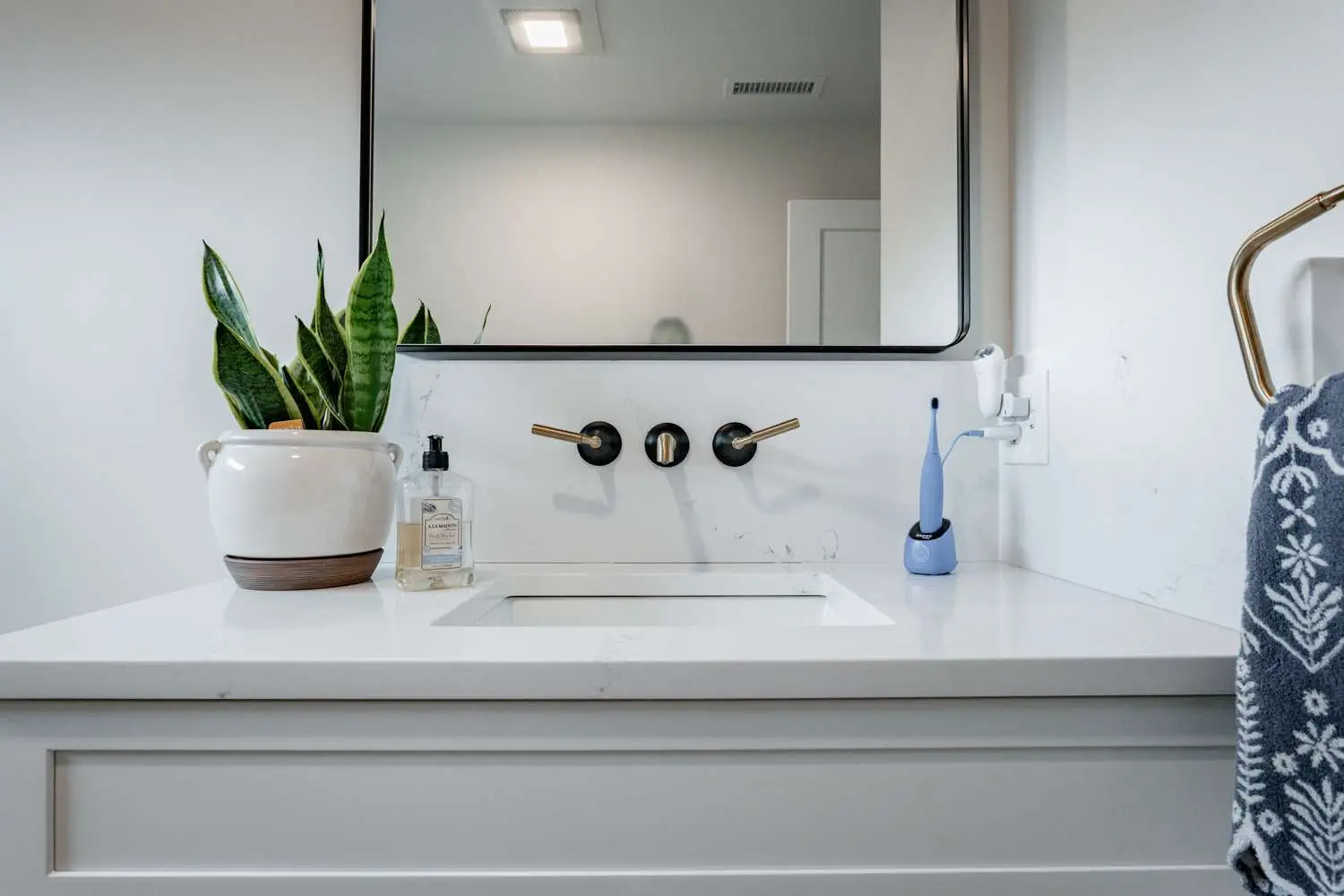 Modern bathroom sink with gold faucet handles, white countertop, potted plant, and personal care items, reflecting Bradford Construction's bathroom remodeling services.