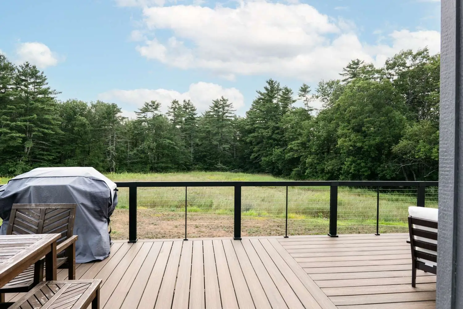 Deck view of custom home in Exeter, NH, featuring wooden patio furniture, a covered grill, and expansive farmland surrounded by trees under a blue sky.