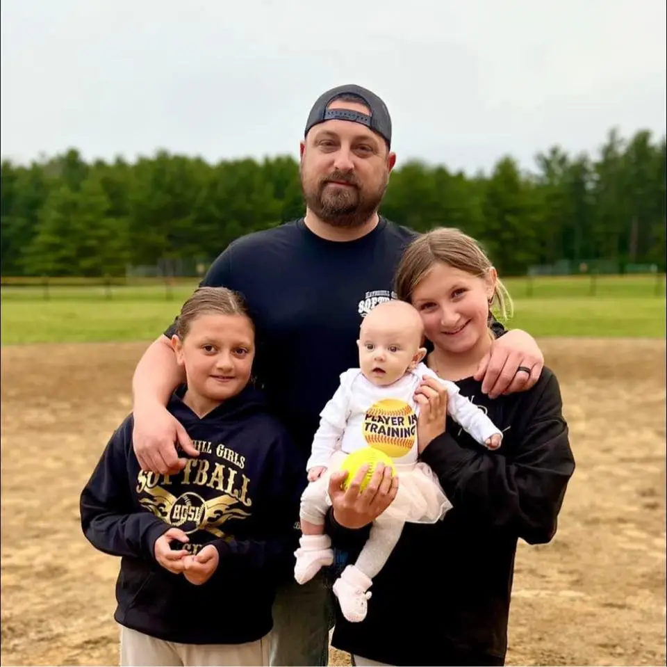 Family portrait of a father with two daughters and a baby, holding a softball, set in a field, reflecting family values and community engagement.