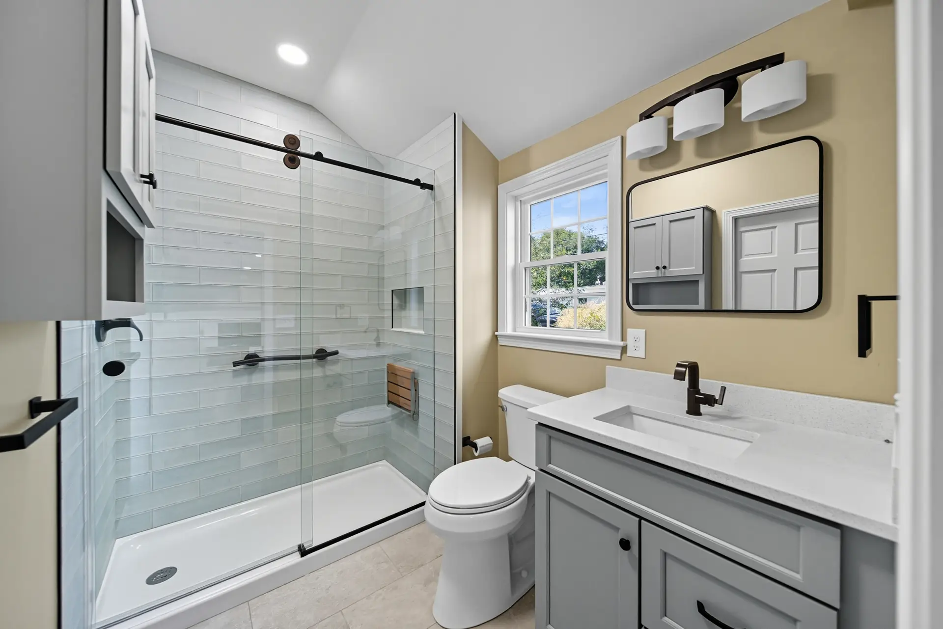 Modern bathroom renovation featuring a glass shower enclosure, gray cabinetry, sleek fixtures, and natural light from a window, emphasizing functional and durable design.