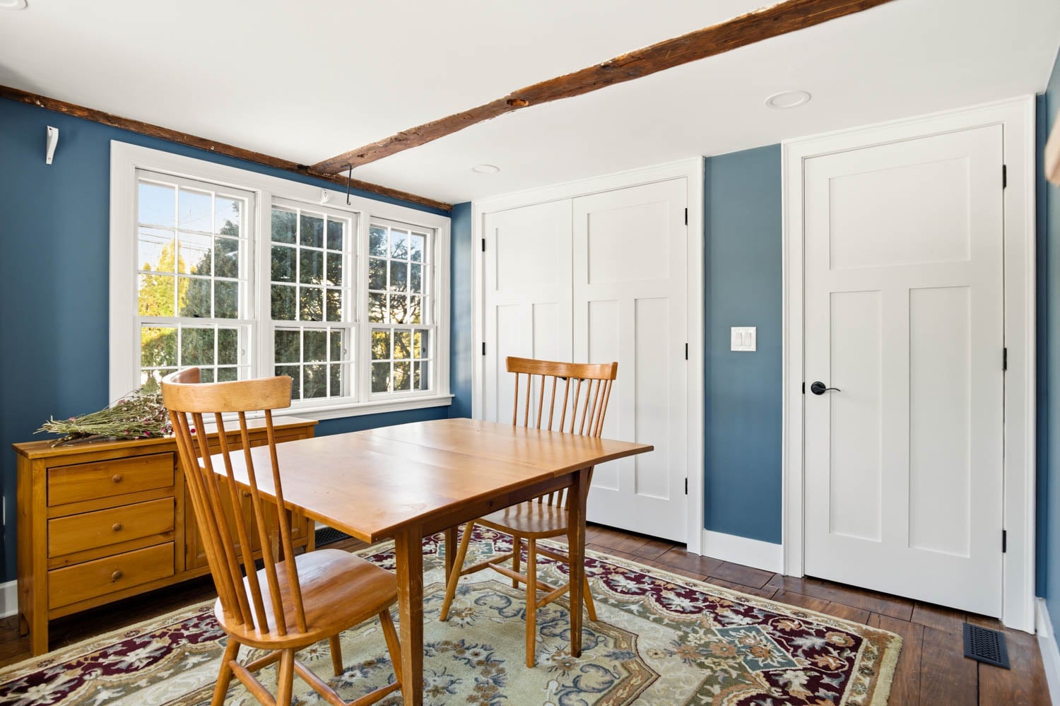 Cozy dining area featuring a wooden table and chairs, with blue walls, natural light from large windows, and a decorative rug, reflecting BCM's home renovation expertise.