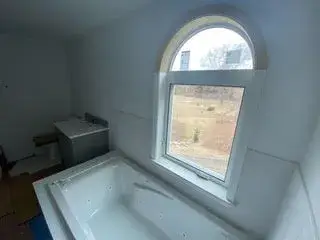 Freestanding soaking tub beneath arched window in modern bathroom of Essex, MA ADU, featuring natural light and serene views.