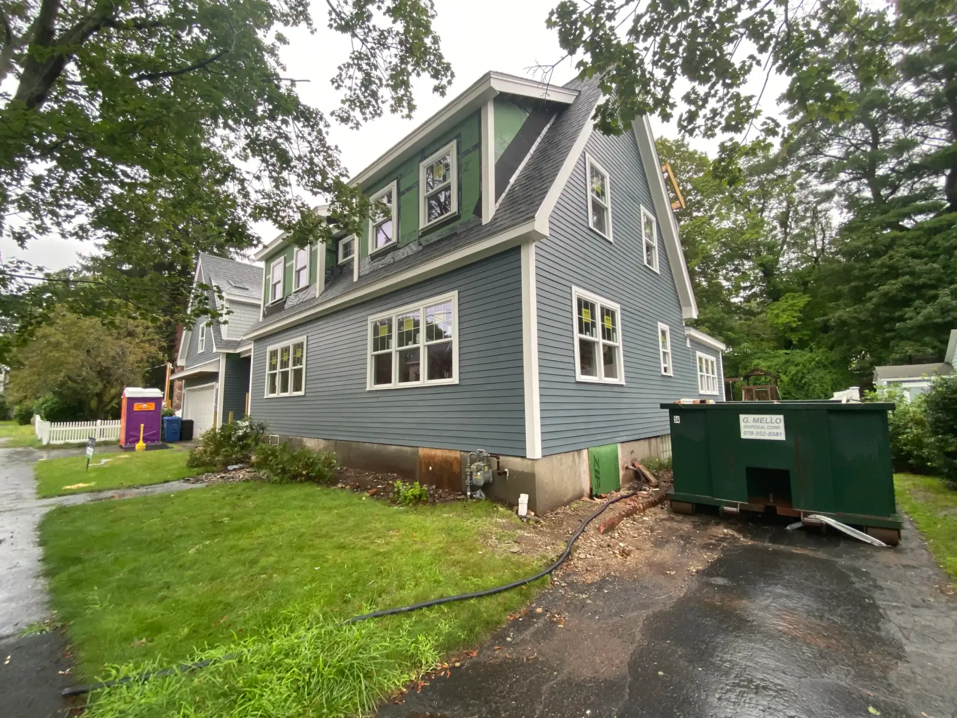 Custom home exterior in Wakefield, MA featuring blue clapboard siding, multiple windows, and a green dumpster, surrounded by landscaping and a residential setting.