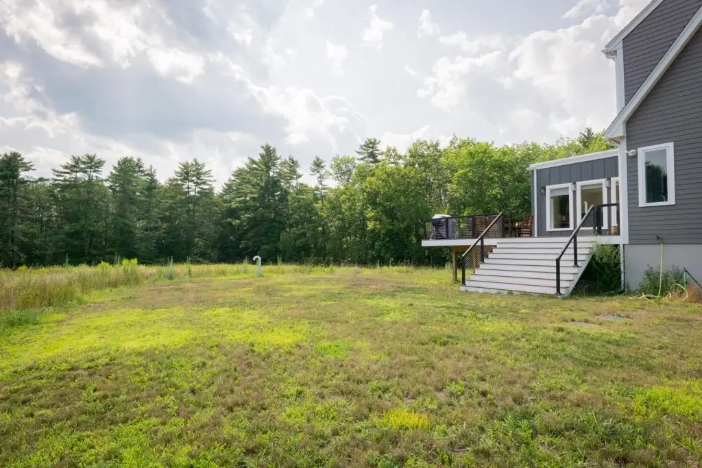 Modern gray house with deck, stairs, and a large grassy backyard with trees.
