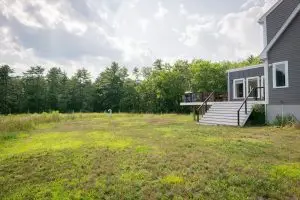 Modern gray house with deck, stairs, and a large grassy backyard with trees.