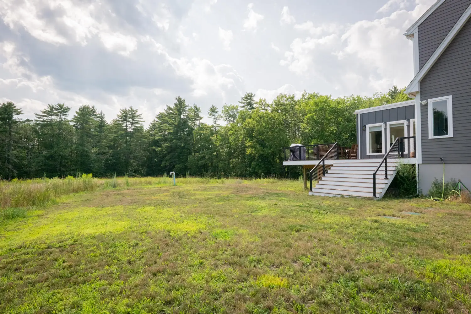 Modern gray house with deck, stairs, and a large grassy backyard with trees.