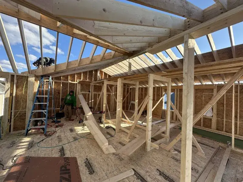 Construction site interior showing framing for a garage-to-ADU conversion in Massachusetts, featuring a ladder, workers, and wooden beams under a blue sky.